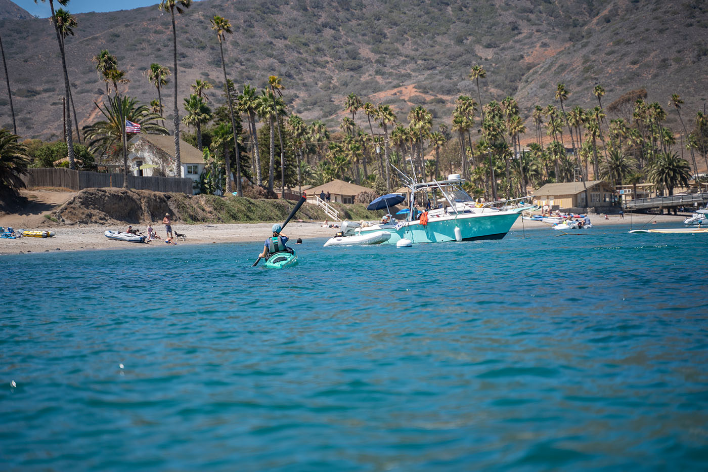 Teal kayak entering Two Harbors past anchored boats and palm trees