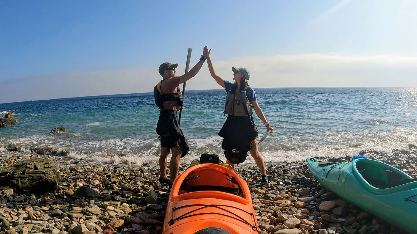 Two kayakers high-five beside their orange and teal Pakayaks on the beach