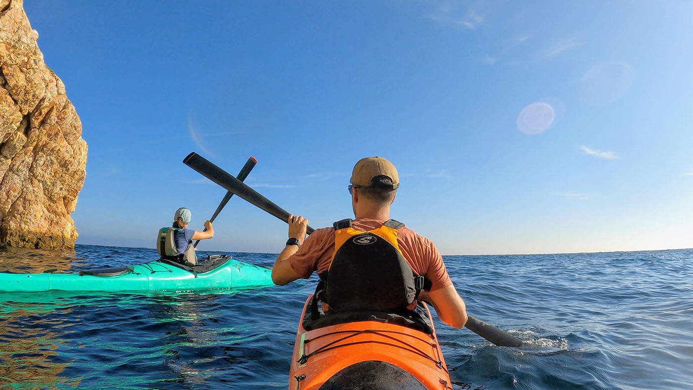 Two kayakers paddling close to a sunlit Catalina Island cliff