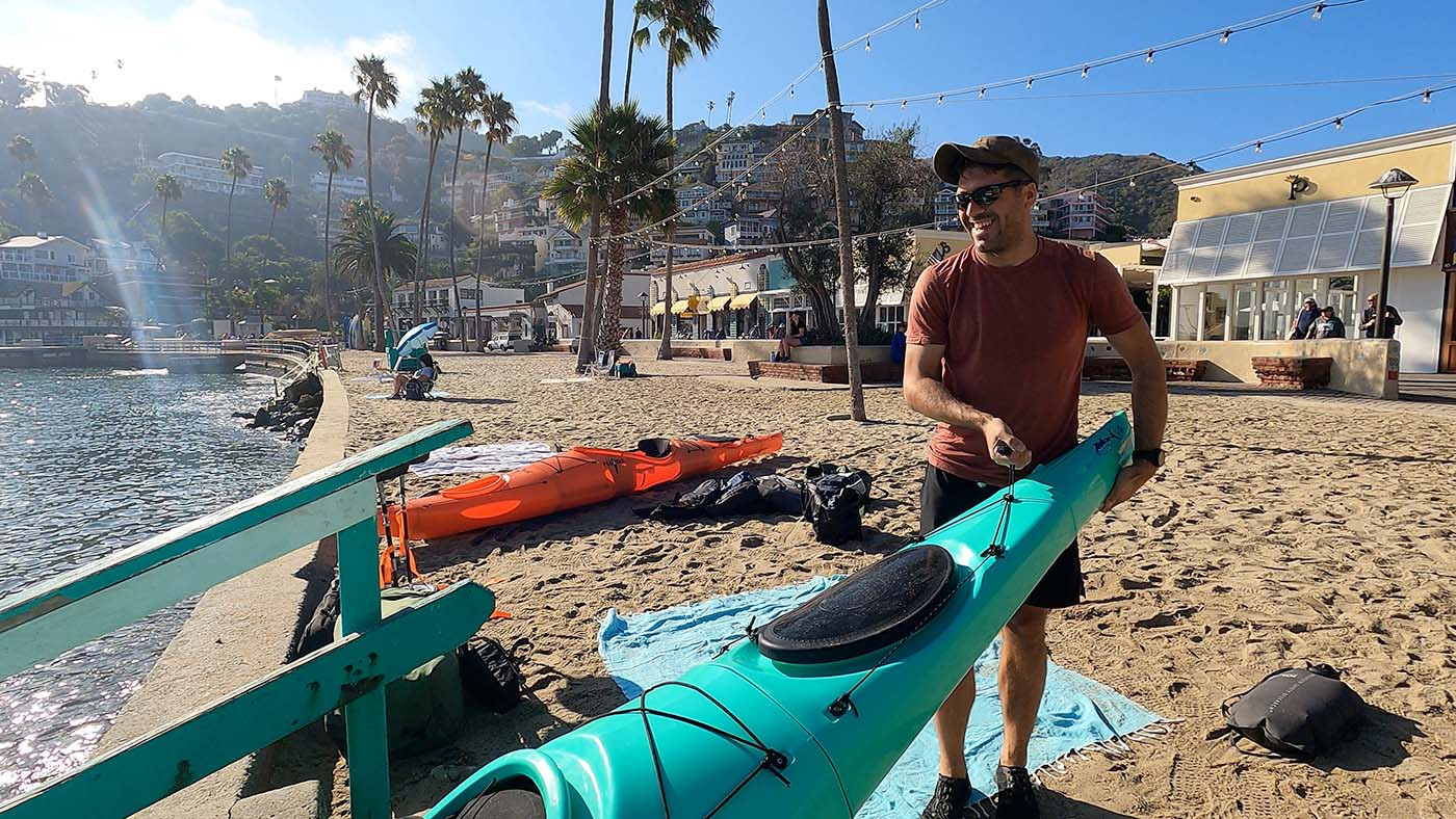 Kayaker carrying an assembled teal Pakayak toward the water in Avalon