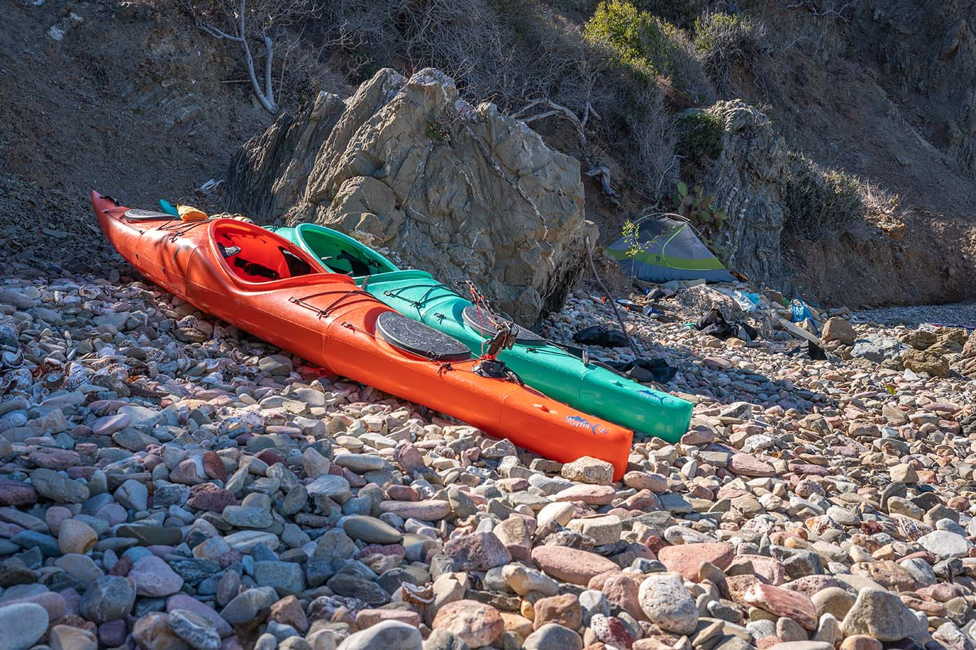 Orange and teal touring kayaks beside a tent on a rocky Catalina Island beach